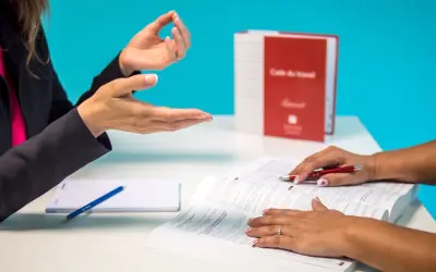 Dwie kobiety rozmawiają przy biurku, jedna wskazuje na otwartą książkę.
