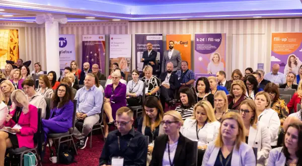 Sala konferencyjna pełna ludzi siedzących w rzędach, z banerami sponsorów w tle.