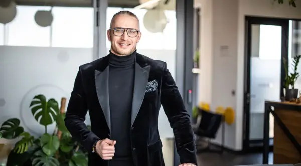 Mężczyzna w czarnym garniturze i okularach stoi w nowoczesnym biurze, uśmiecha się.