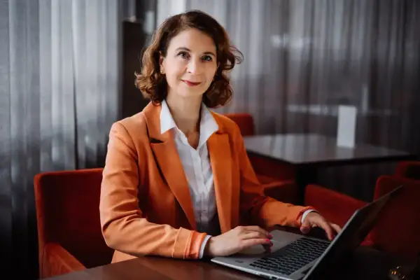 Kobieta w pomarańczowej marynarce pracuje na laptopie w eleganckim wnętrzu.